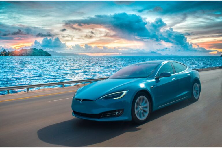 A blue tesla car moving along the road with a sea view at the back.