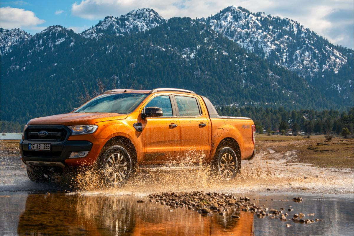 Side view image of Ford Ranger in orange color driving in the mud with mountain background.