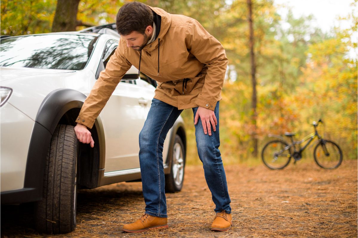 A photo of man bending his knees, checking the tyre of white car.