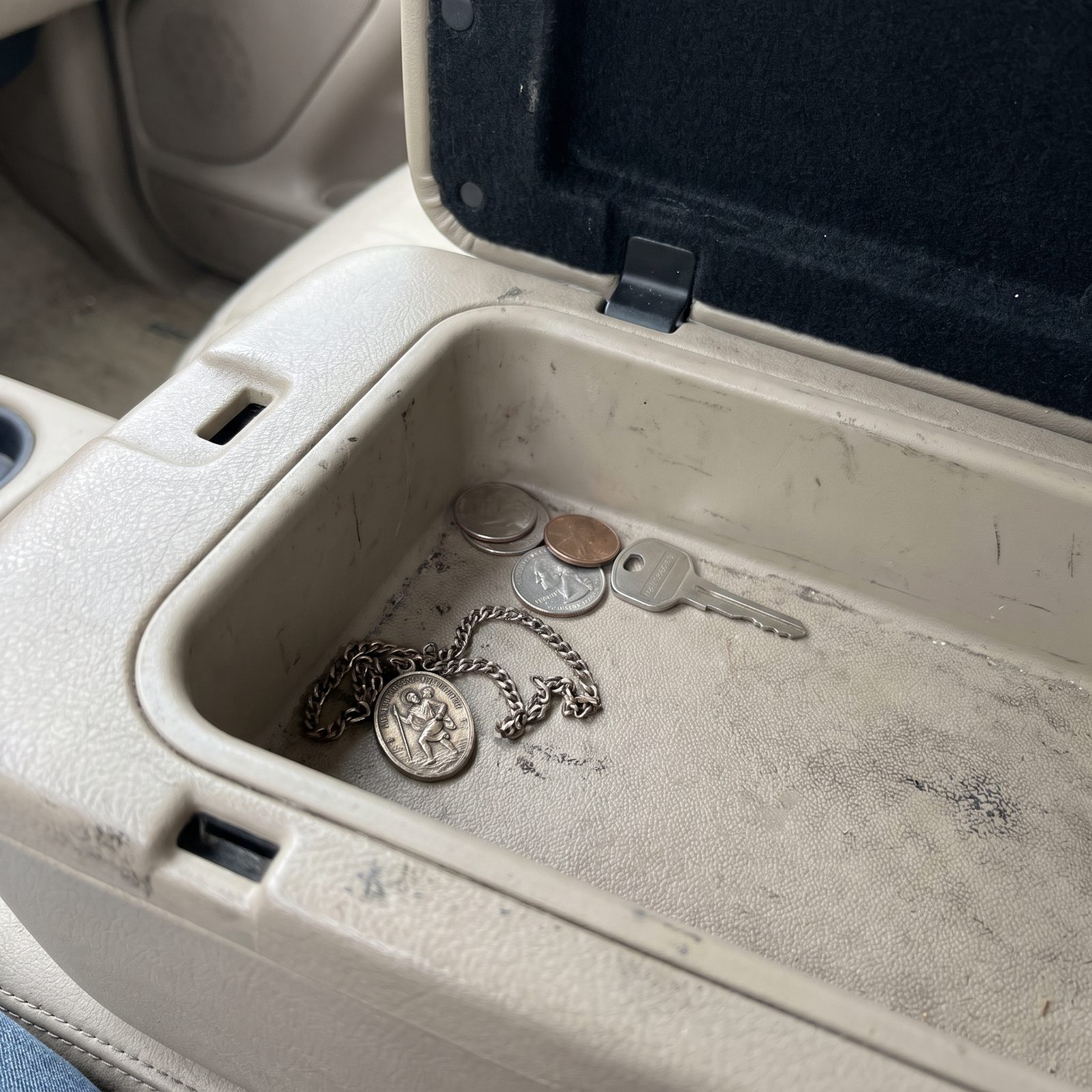 A small worn St. Christopher medal resting alone in the corner of an open car console tray, with a few coins and a key set