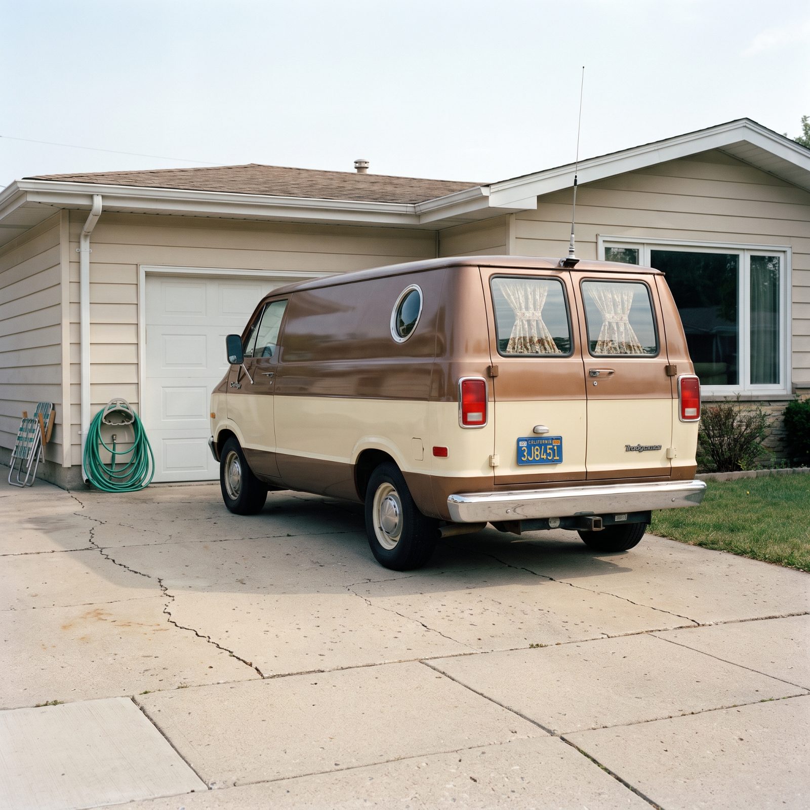 Suburban driveway with a brown-and-cream 1977 custom van showing a round side window and curtains in the rear glass.