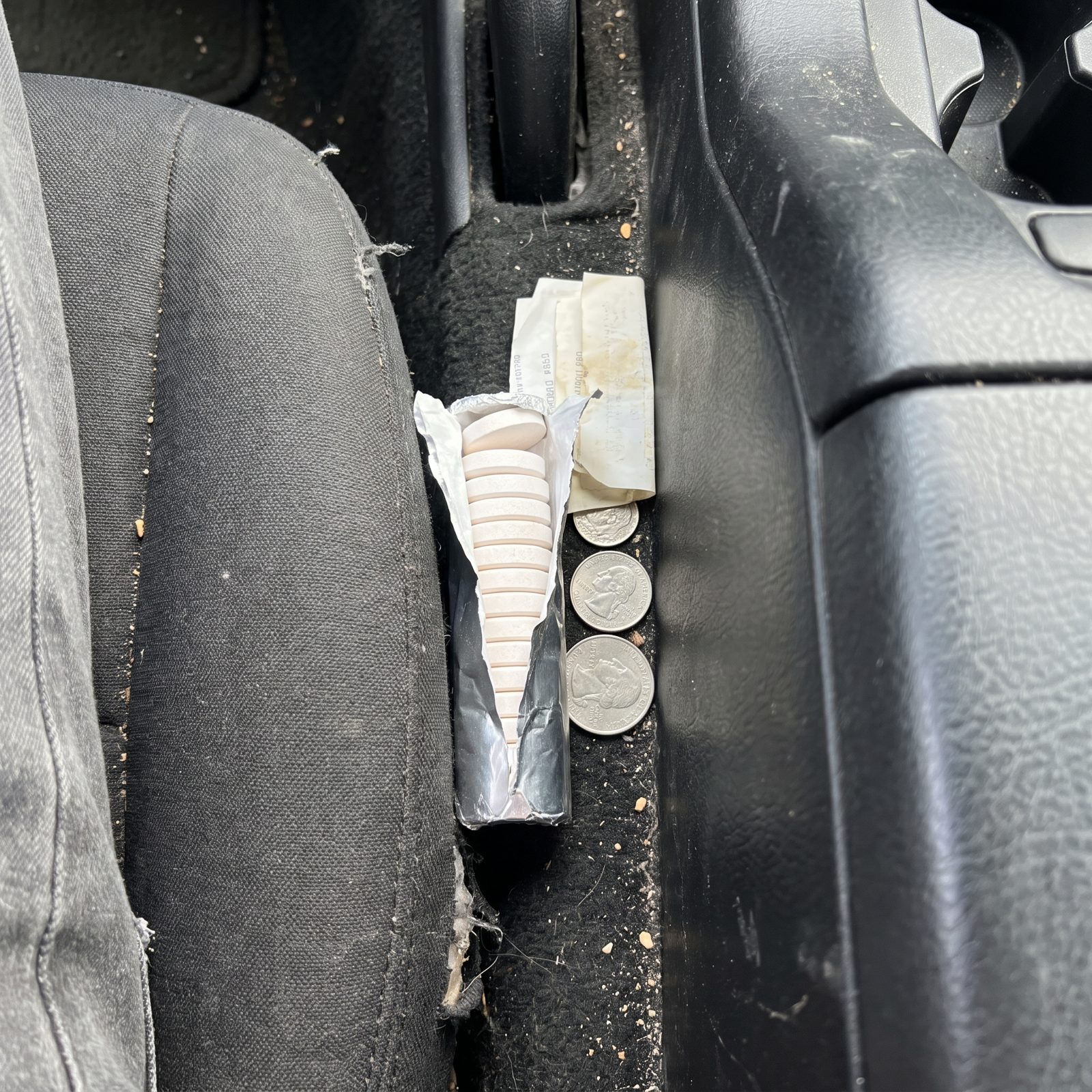 A half-used roll of antacid tablets wedged into the seam between a car seat and center console, with loose coins and a