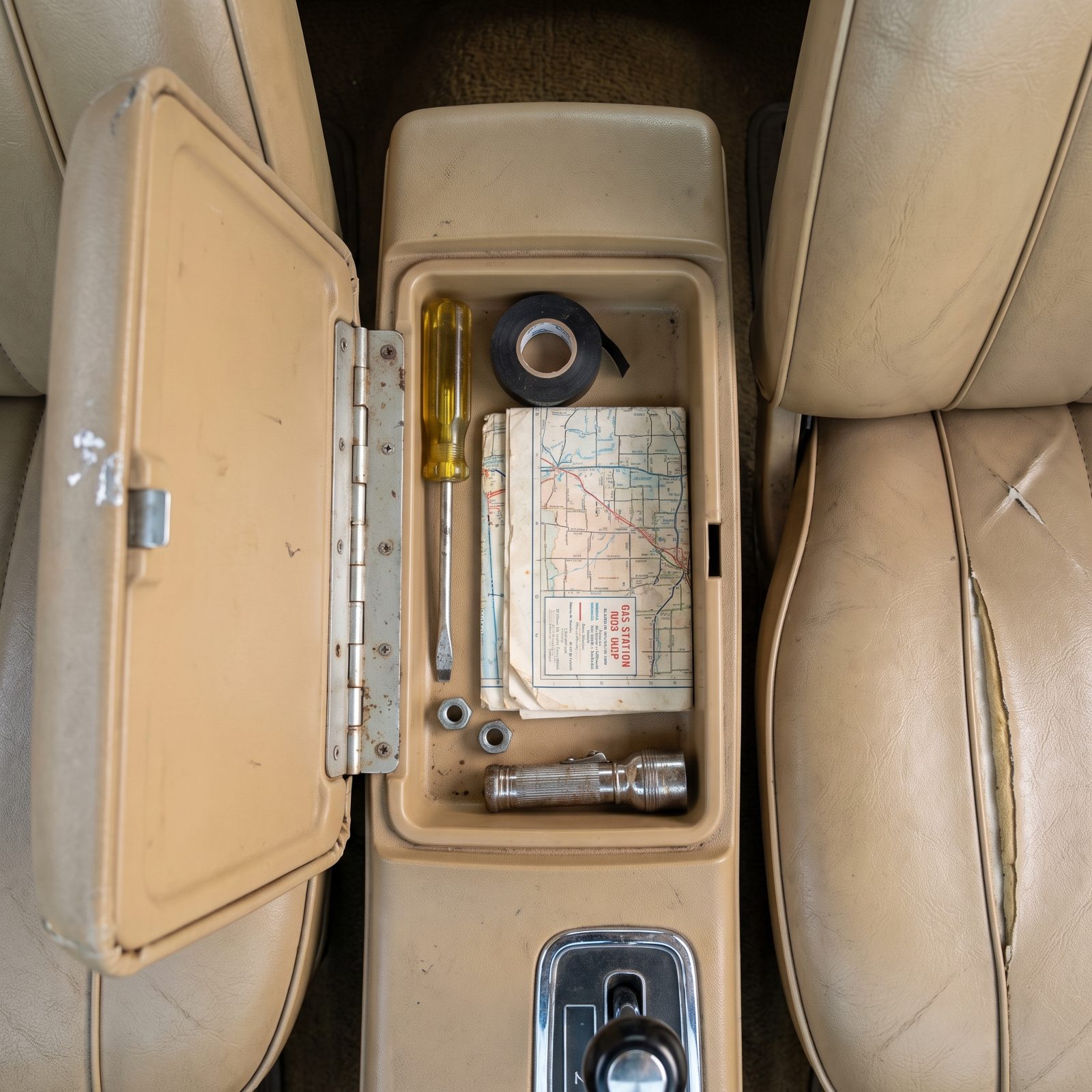 Open center console tray in a 1970s sedan holding a yellow-handled screwdriver, electrical tape, a folded road map, hex