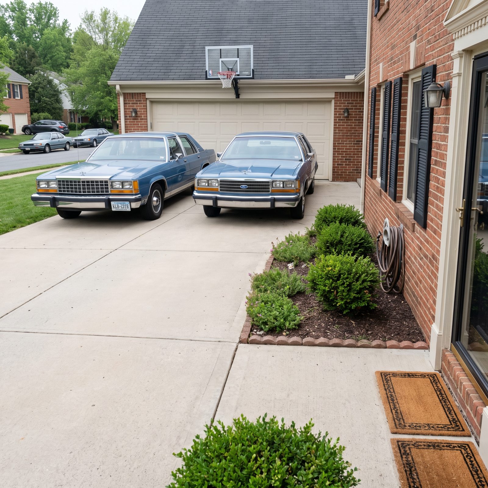Two identical medium-blue 1980s sedans parked side by side facing the same way in a two-car driveway.