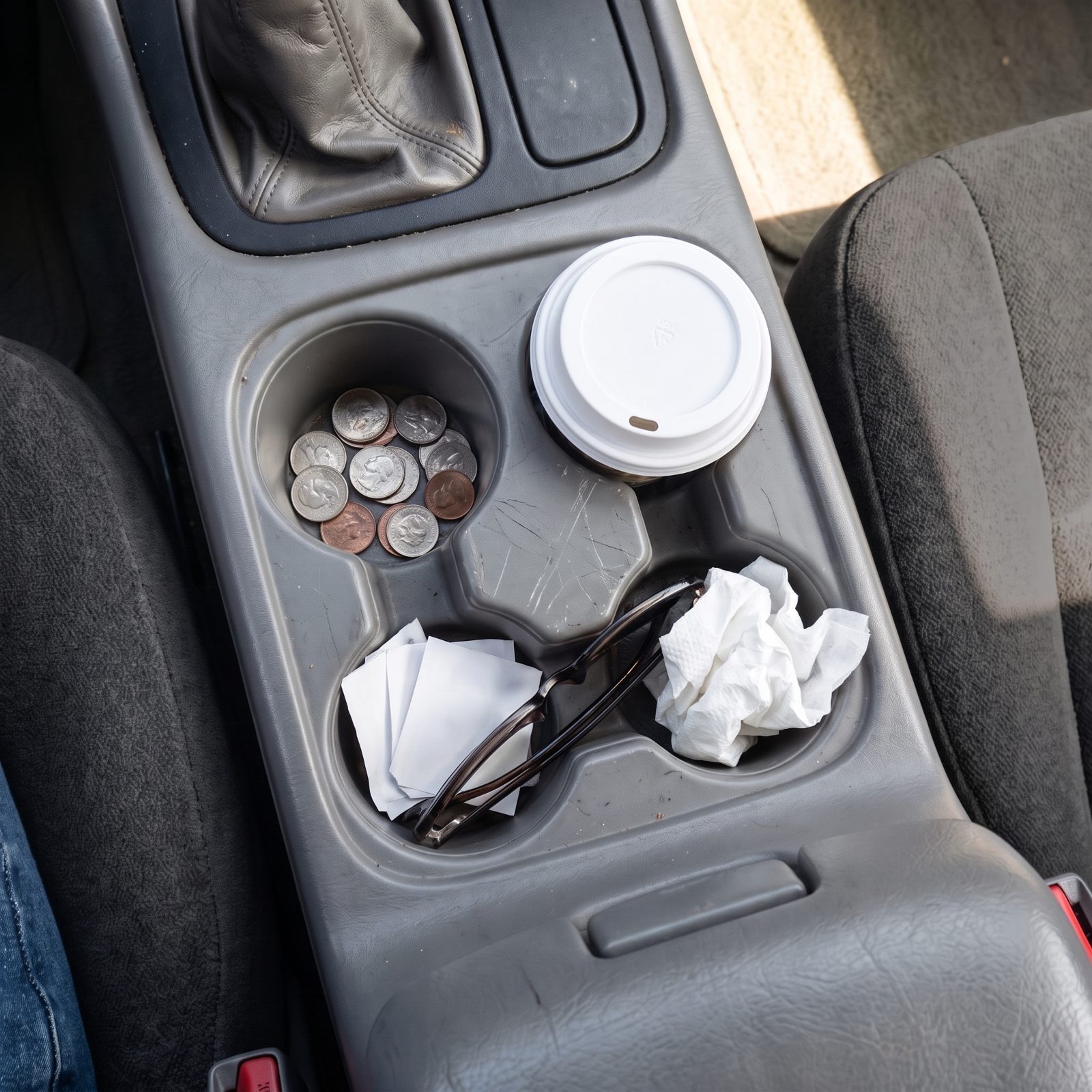 Center console of a 1990s car from above holding two coffee cup lids, loose coins, three parking stubs, sunglasses, and one