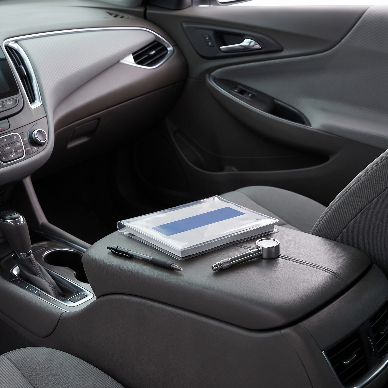 Open center console holding a car owner's manual in a vinyl sleeve on top of folded receipts, with a pen and a tire-pressure