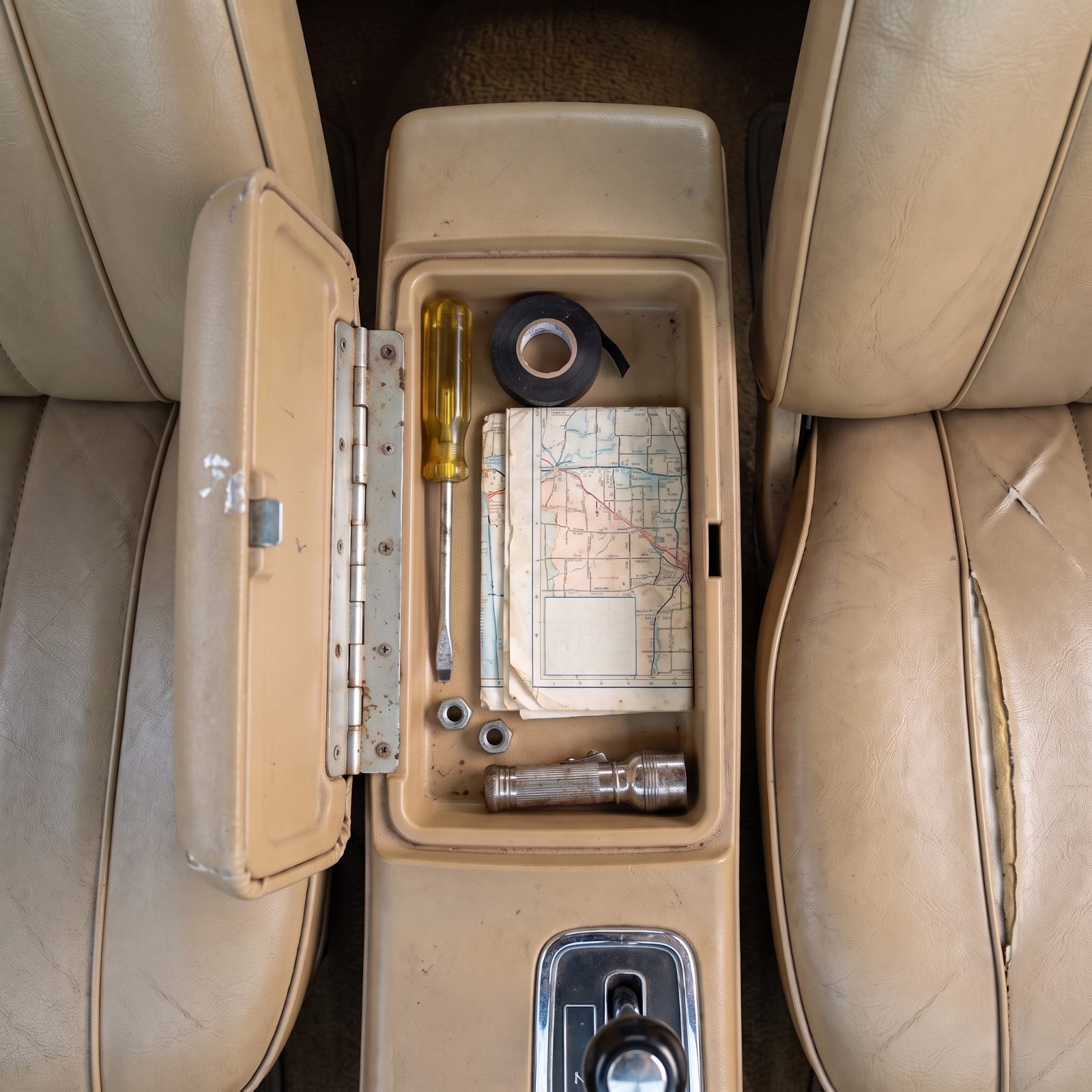 Open center console tray in a 1970s sedan holding a yellow-handled screwdriver, electrical tape, a folded road map, hex
